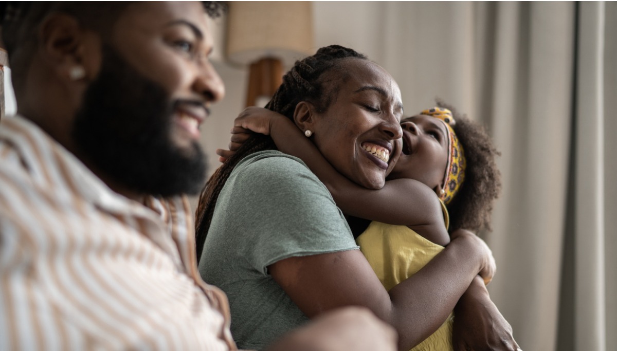 A family sitting in a living room and a daughter has her arms wrapped around her mother's neck