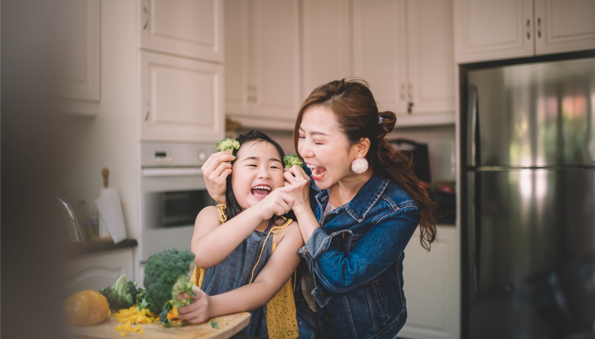 A mother and child laughing and eating broccoli in a kitchen