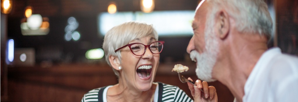 A older woman feeding food to her husband and laughing