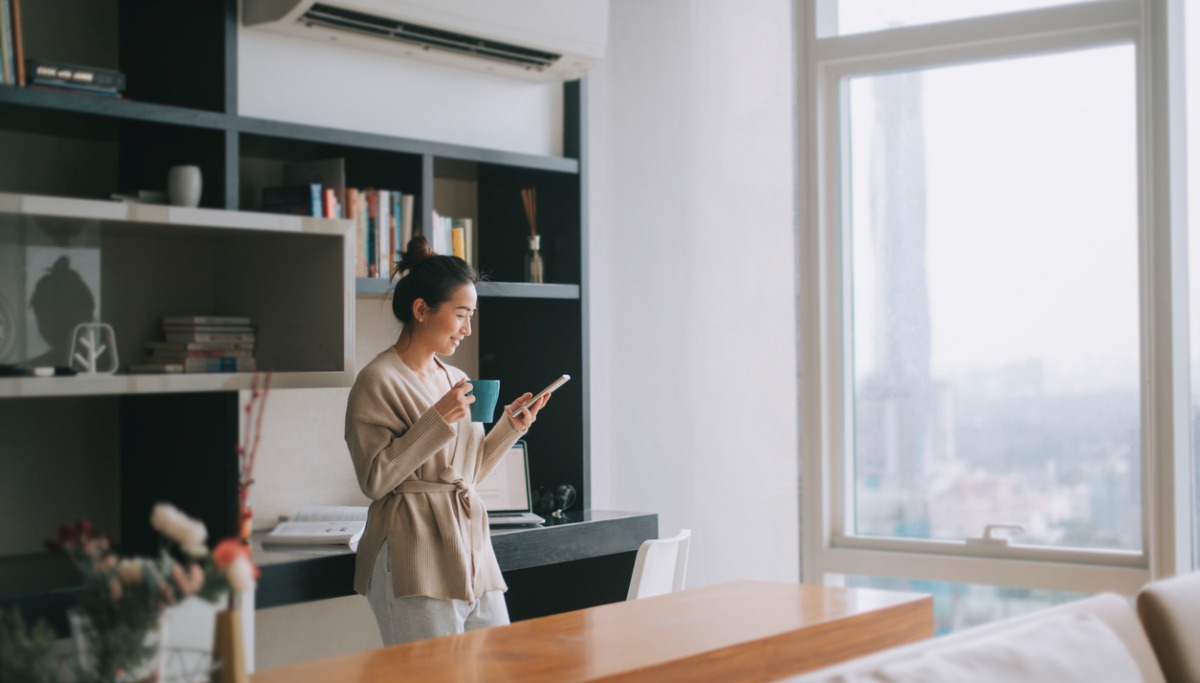 A woman standing in a sky rise condo and looking at her phone while holding a cup