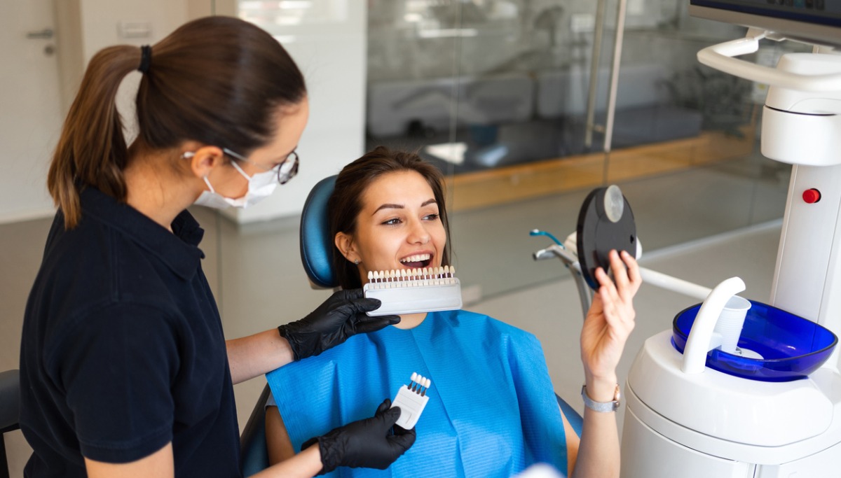 A patient looking in a mirrow as a dentist shows different colors of veneers