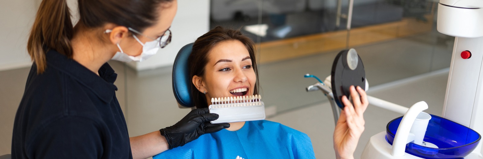 A patient looking in a mirrow as a dentist shows different colors of veneers