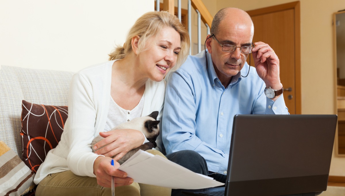 An older couple sitting on a couch with a cat and looking at a laptop together