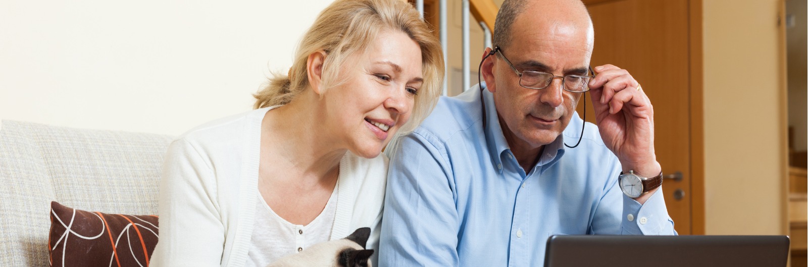 An older couple sitting on a couch with a cat and looking at a laptop together
